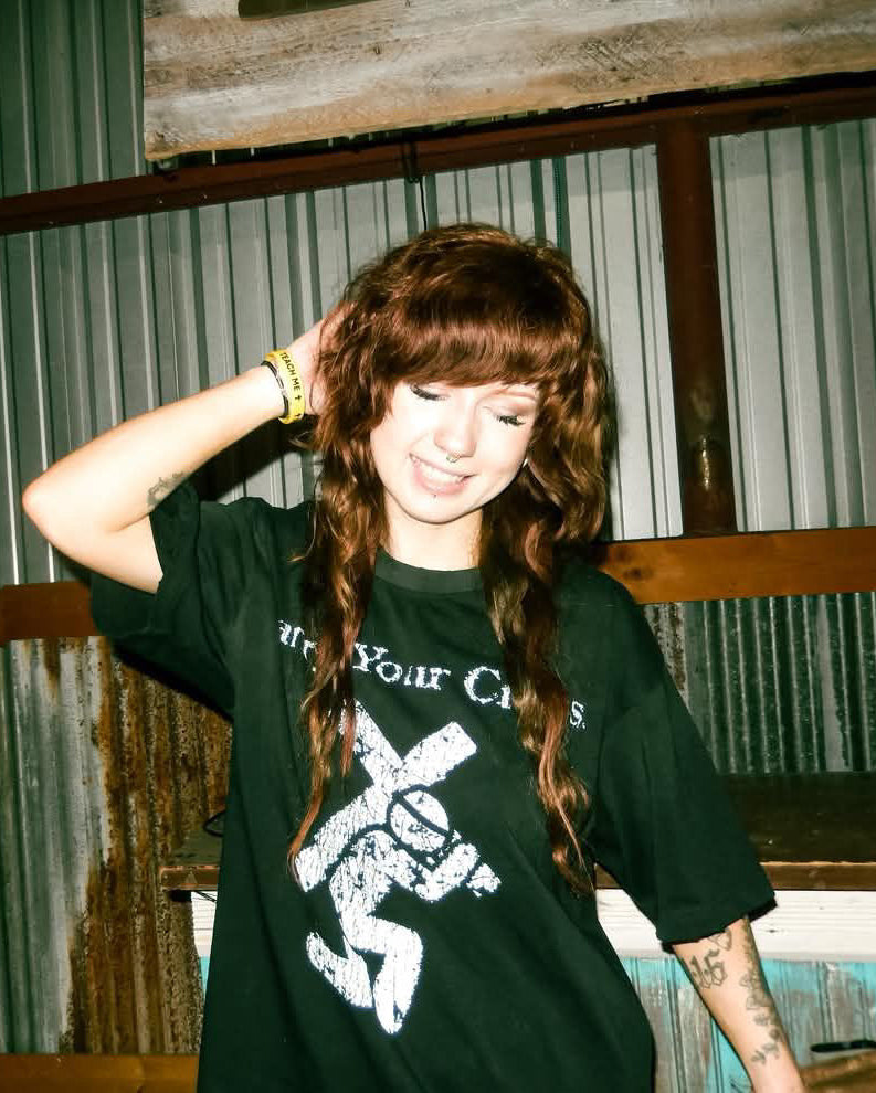 A woman with long wavy brown hair wearing a black graphic t-shirt with a white distressed print, posing in front of a corrugated metal wall.