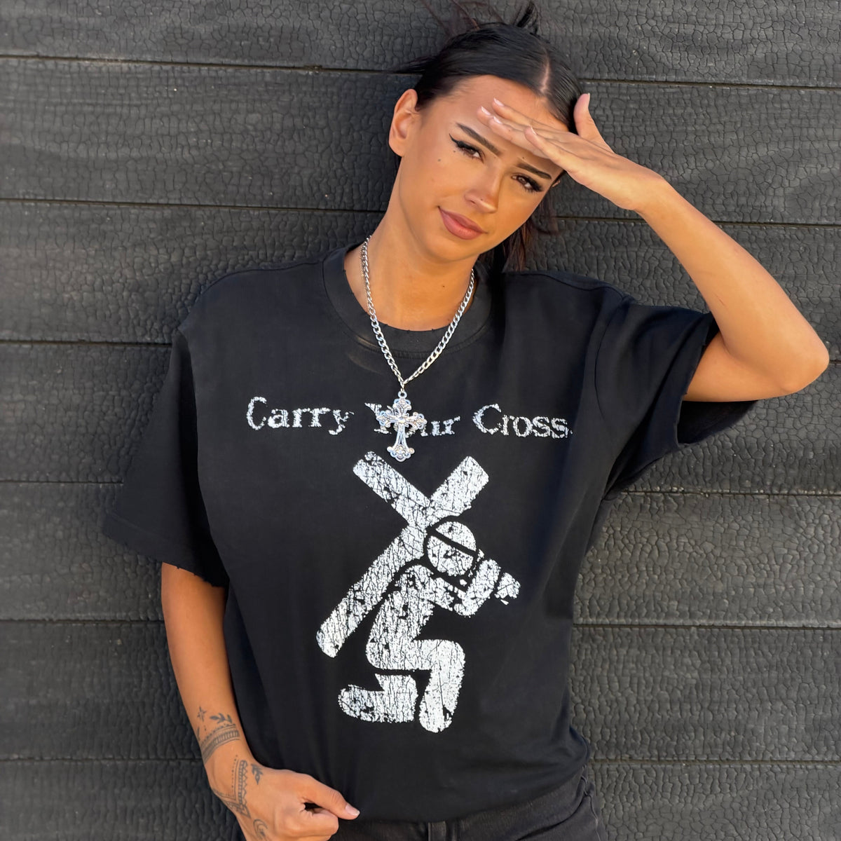A woman wearing a black graphic t-shirt with a distressed white print that reads 'Carry Your Cross' and shows a cross-shaped figure. She is posing against a dark gray wooden plank wall.